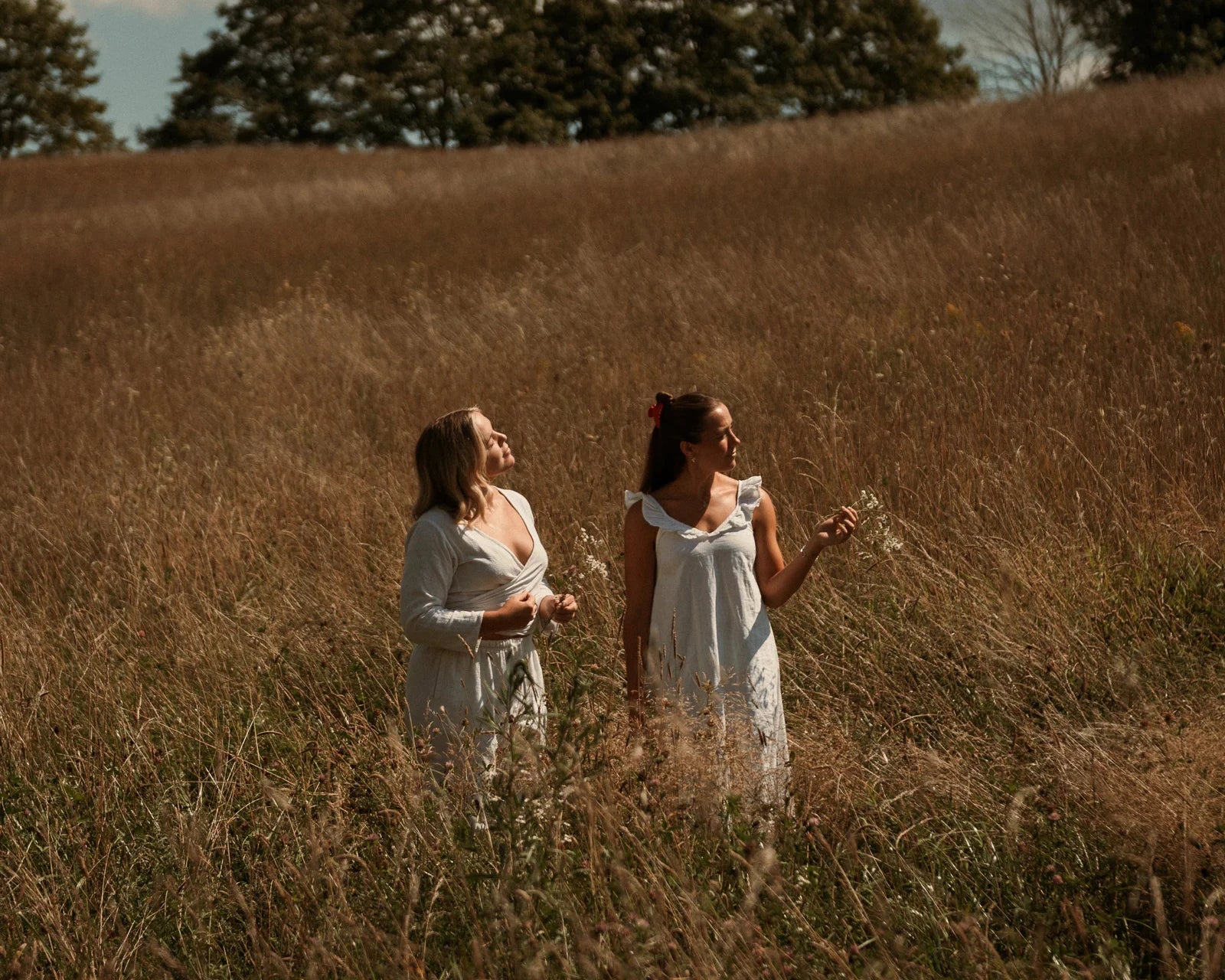 Two women in white dresses standing in a field of tall grass with trees in the background